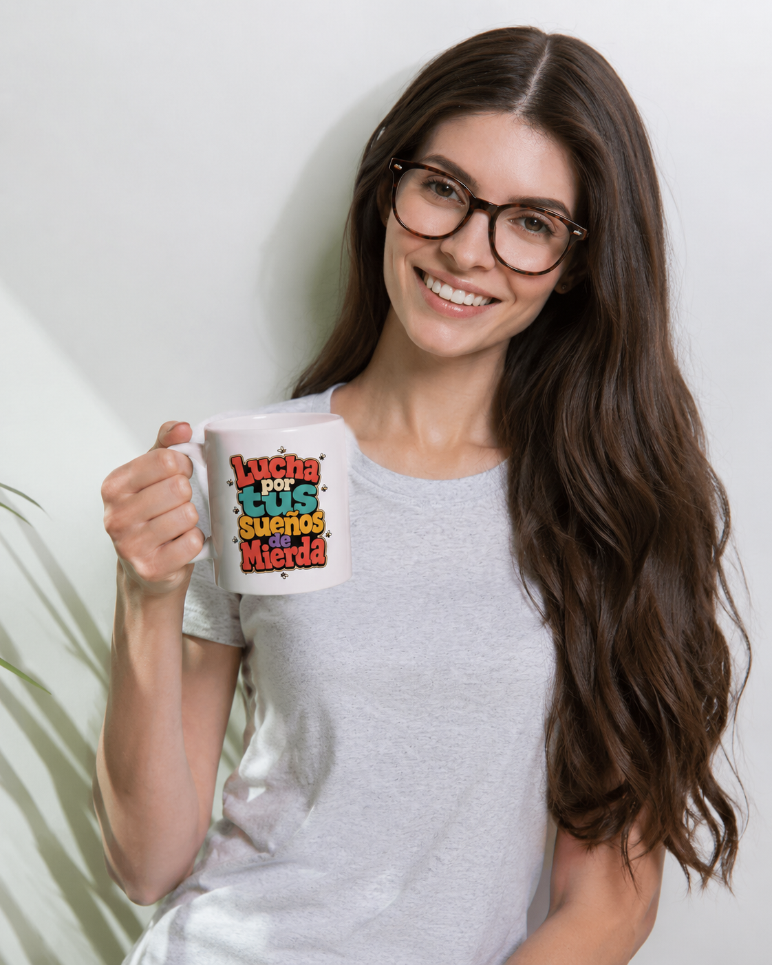 A smiling woman with long brown hair and glasses holds up a white ceramic mug with a colorful graphic that reads 'Lucha Luz Sueno Mierda' in red, green, yellow, and blue text. She is wearing a light gray t-shirt against a white wall.