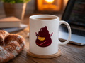 A white ceramic coffee mug with a cartoon poop character wearing a ninja outfit, sitting on a wooden coaster on a wooden desk with a laptop, a knit blanket, and a warm light in the background.